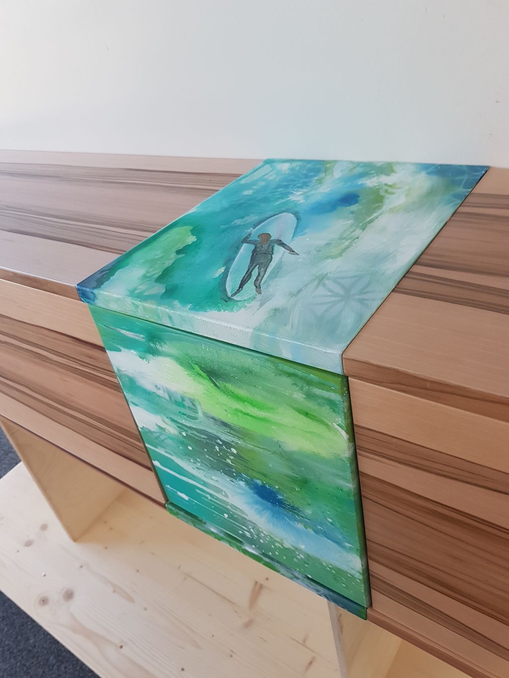 Ein Gemälde mit einem Surfer auf einem Surfbrett in blau-grünen Wassern, das an den Kanten herunterhängt, und auf einer braunen Holzoberfläche liegt.
