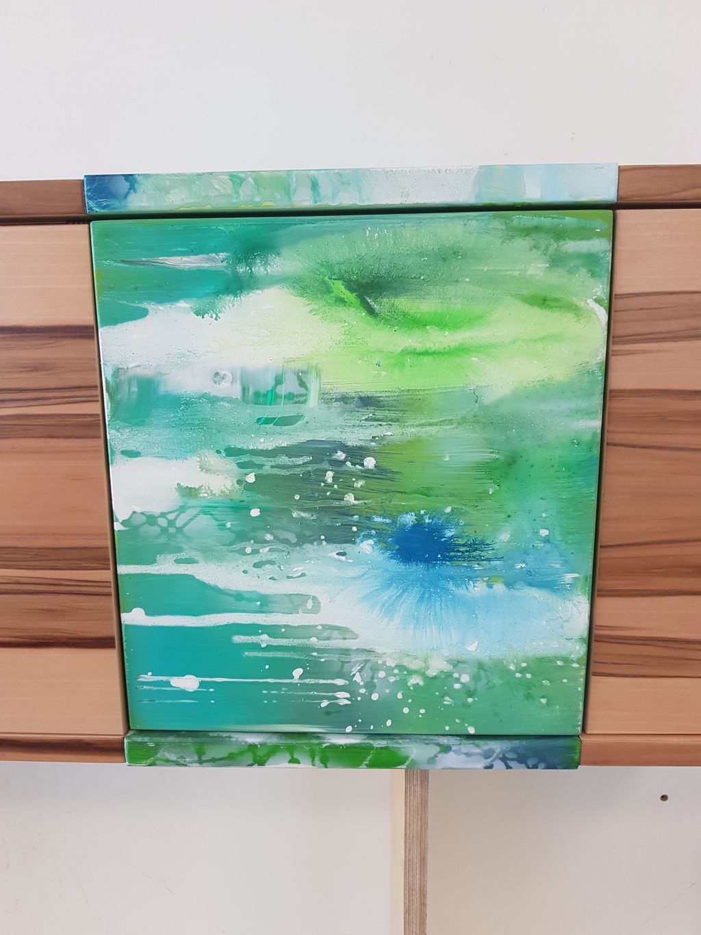 Abstraktes Gemälde mit dominierenden Grüntönen, blau-weißen und türkisen Farbspritzern, in einem Holzrahmen an einer weißen Wand.